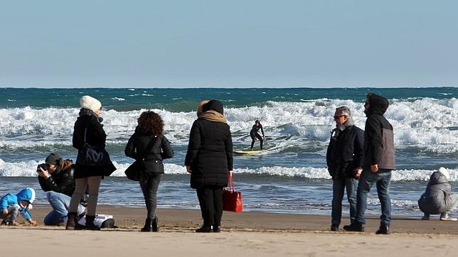 Un hombre practica surf mientras varias personas pasean por la orilla