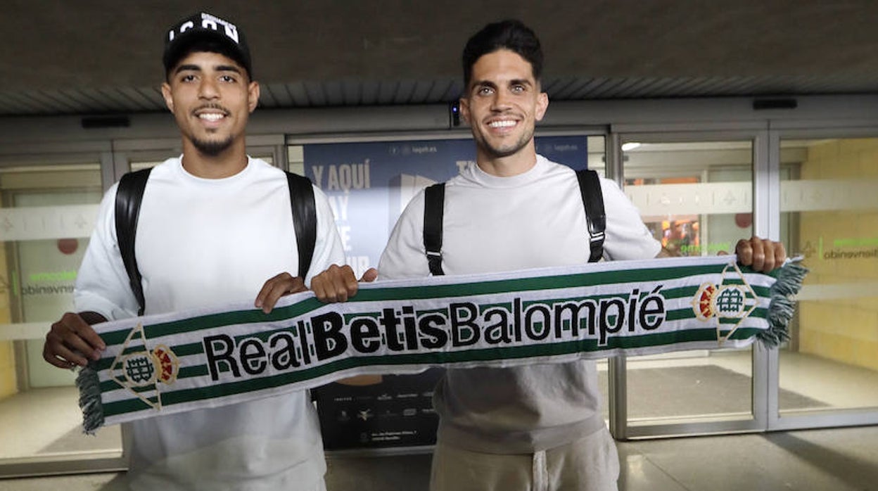 Chadi Riad y Marc Bartra posan con una bufanda del Betis en una de las puertas de la terminal de llegadas del Aeropuerto de Sevilla