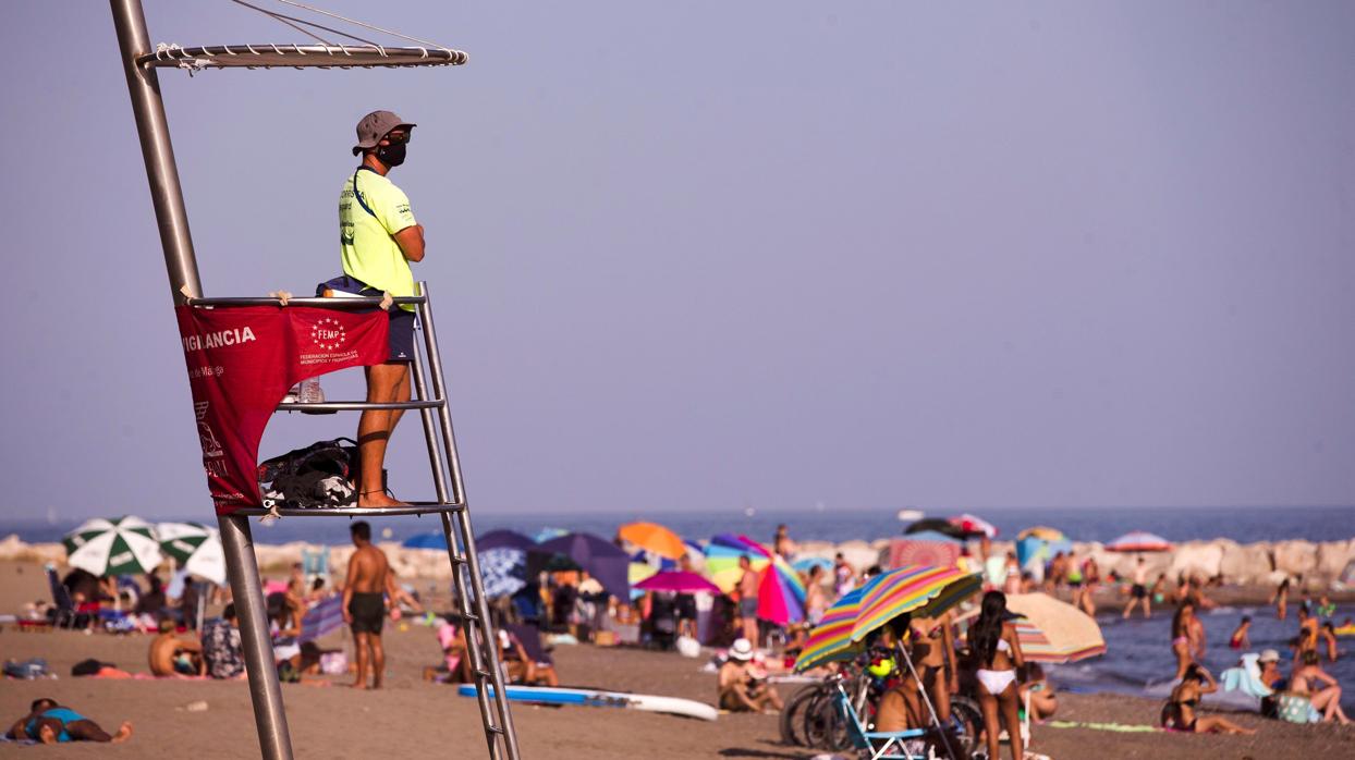 Un vigilante con mascarilla observa a los bañistas de la playa de la Malagueta