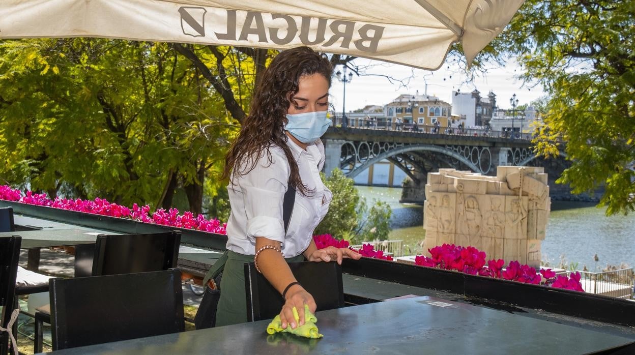 Una camarera limpia una mesa en la terraza de la lonja del Barranco de Sevilla con vistas al puente de Triana