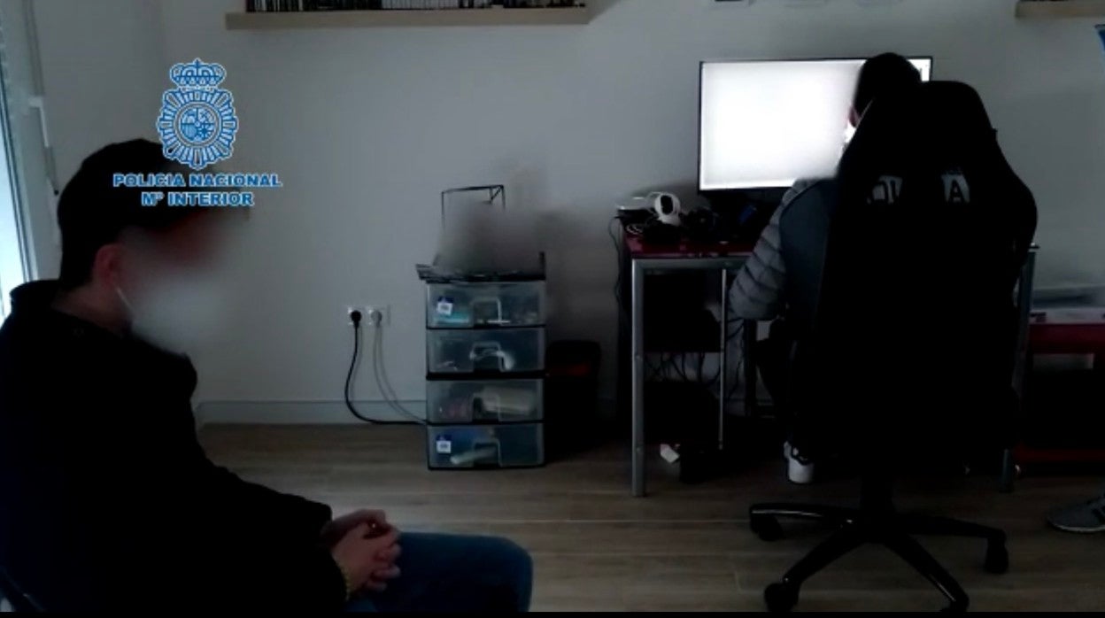 El detenido en Sevilla sentado en una silla mientras un agente inspecciona sus equipos informáticos