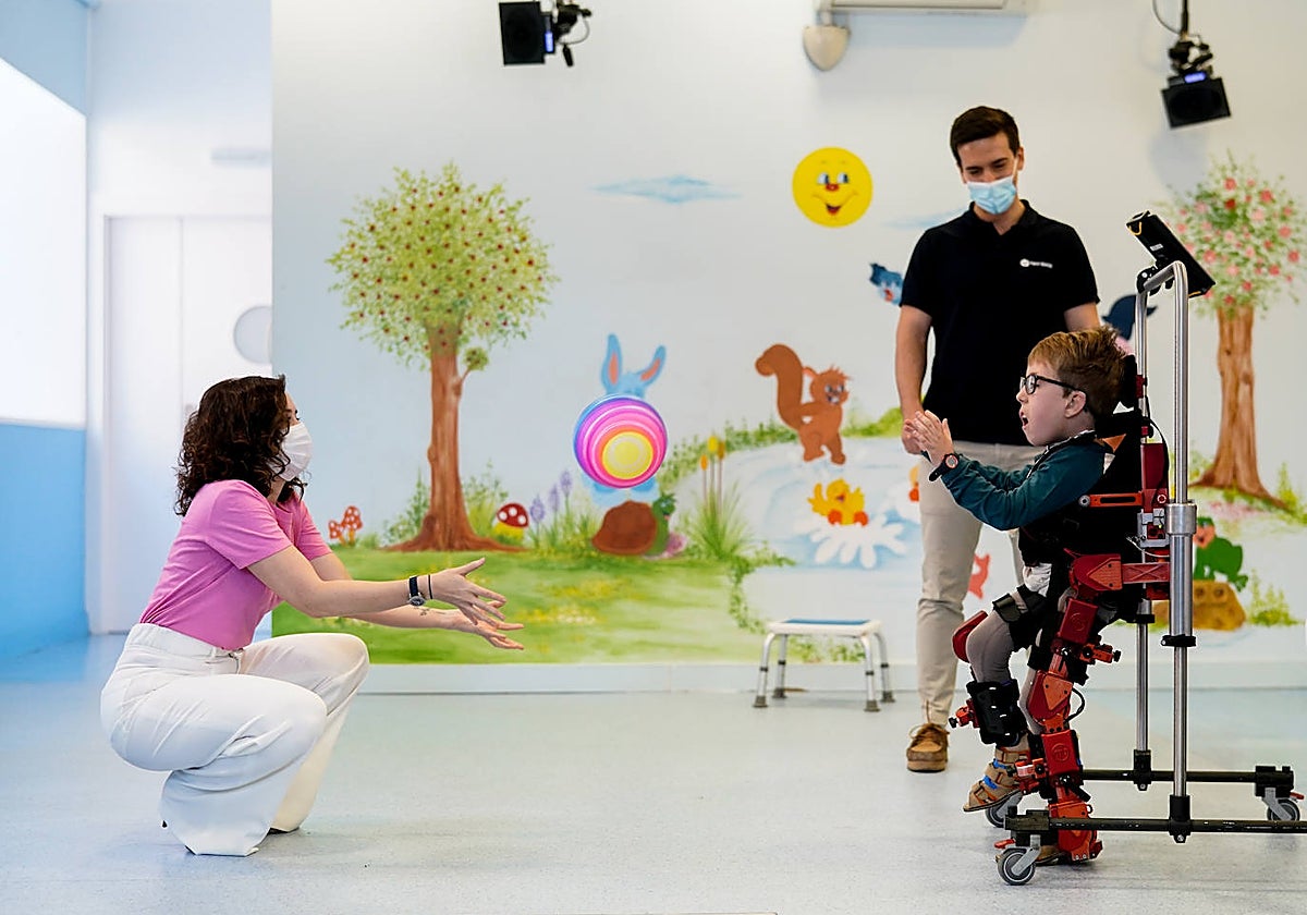 Adolfo, en el exoesqueleto, juega con una pelota con la presidenta Díaz Ayuso, bajo la mirada del técnico del aparato, Fernando