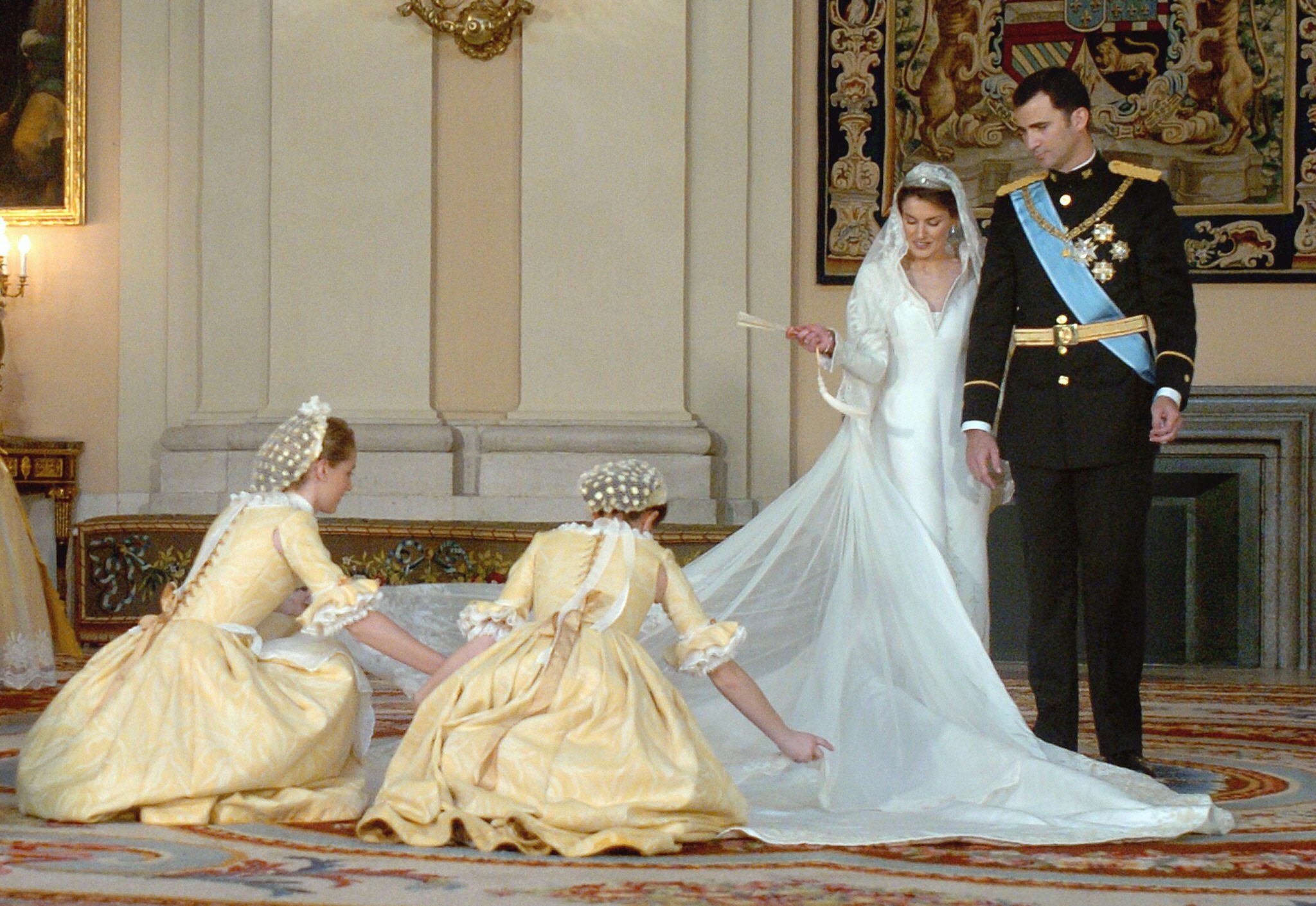 Don Felipe y Doña Letizia aguardan mientras las dos damas de honor que compusieron el cortejo nupcial de la Reina, Ana Codorniú y Álvarez de Toledo, hija de Victoria Eugenia Álvarez de Toledo, marquesa de Casa Loring, y de Alfonso Codorniú Aguilar y, por ende, bisnieta de la infanta María Cristina de Borbón; y Claudia González Ortiz, prima de la entonces Princesa, le arreglan la cola del vestido para la foto oficial con todos los miembros de la familia. Las jóvenes lucieron un vestido del diseñador Lorenzo Caprile de inspiración goyesca. El objetivo del artista era emular los trajes que los encargados de atender a la Familia Real vestían en el siglo XVIII.
