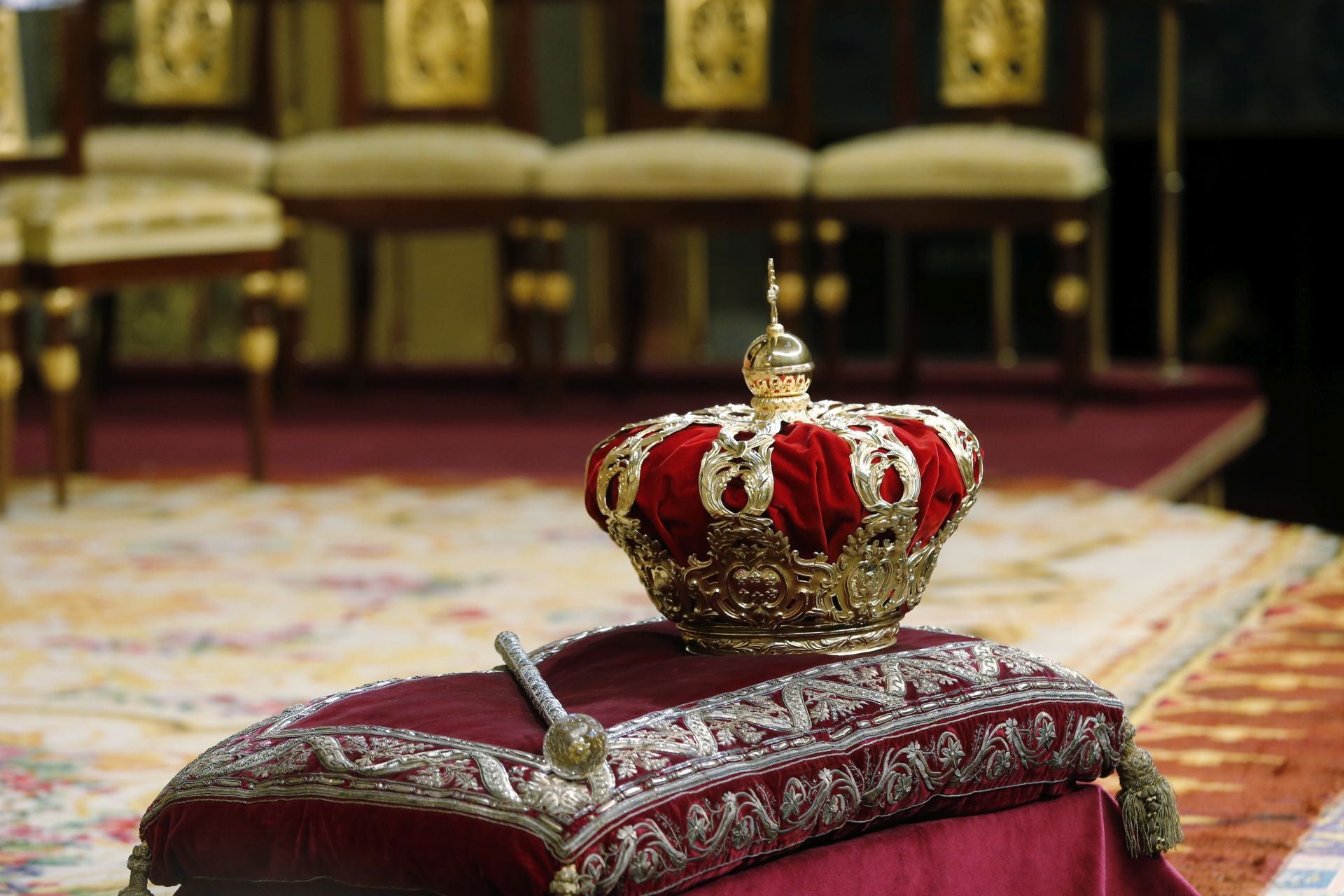 En esta imagen se puede apreciar, sobre un cojín, la Corona y el Cetro reales que presidieron la ceremonia de proclamación de Felipe VI en el Congreso de los Diputados. Estos objetos, símbolos de la máxima representación de la Monarquía española, proceden de los reinados de Carlos II y Carlos III. Desde el reinado de Isabel II, se han utilizado en diversos actos reales, así como en ceremonias fúnebres, siempre con un carácter decorativo. Antes del acto que convirtió a Don Felipe en el nuevo monarca de España, ambos objetos pudieron apreciarse en el traslado de los restos de Alfonso XIII a España para su enterramiento en el monasterio de El Escorial, en 1980. La Corona Real, de estilo neoclásico y formada con emblemas heráldicos y ramas de laurel, fue elaborada por Fernando Velasco, platero de cámara de la Real Casa desde 1748. El encargo, cuyo motivo se desconoce, tuvo lugar durante el reinado de Carlos III. El Cetro Real, un bastón de mando formado por tres cañones de plata dorada, apareció en 1701 registrado en el inventario que se realizó a la muerte de Carlos II, pero se desconoce su origen. Tanto la Corona como el Cetro pertenecen a las Colecciones Reales de Patrimonio Nacional y se encuentran custodiados en la cámara acorazada del Palacio Real de Madrid.