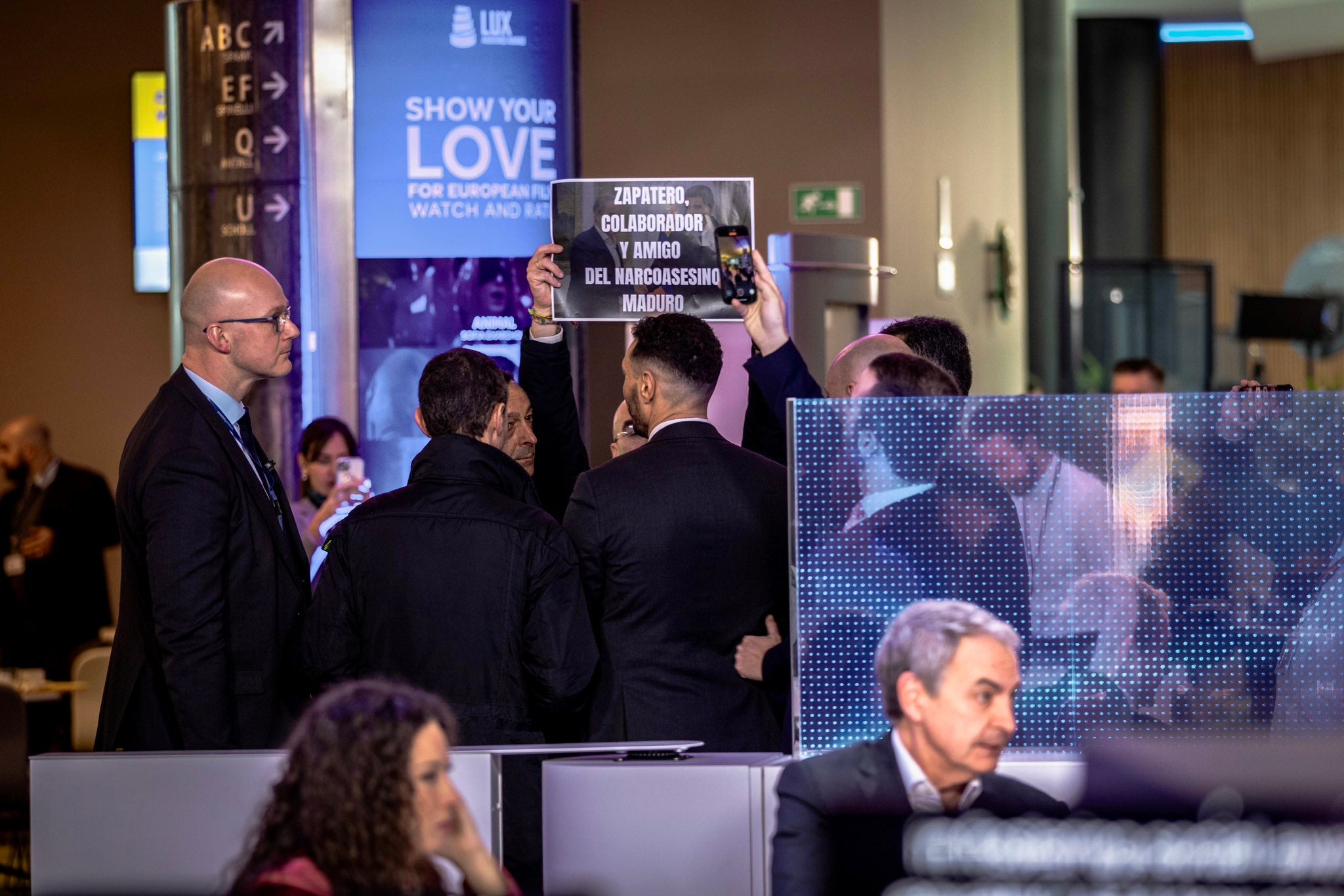 Jorge Buxadé levante un cartel contra Zapatero mientras el expresidente participa en un acto en Bruselas