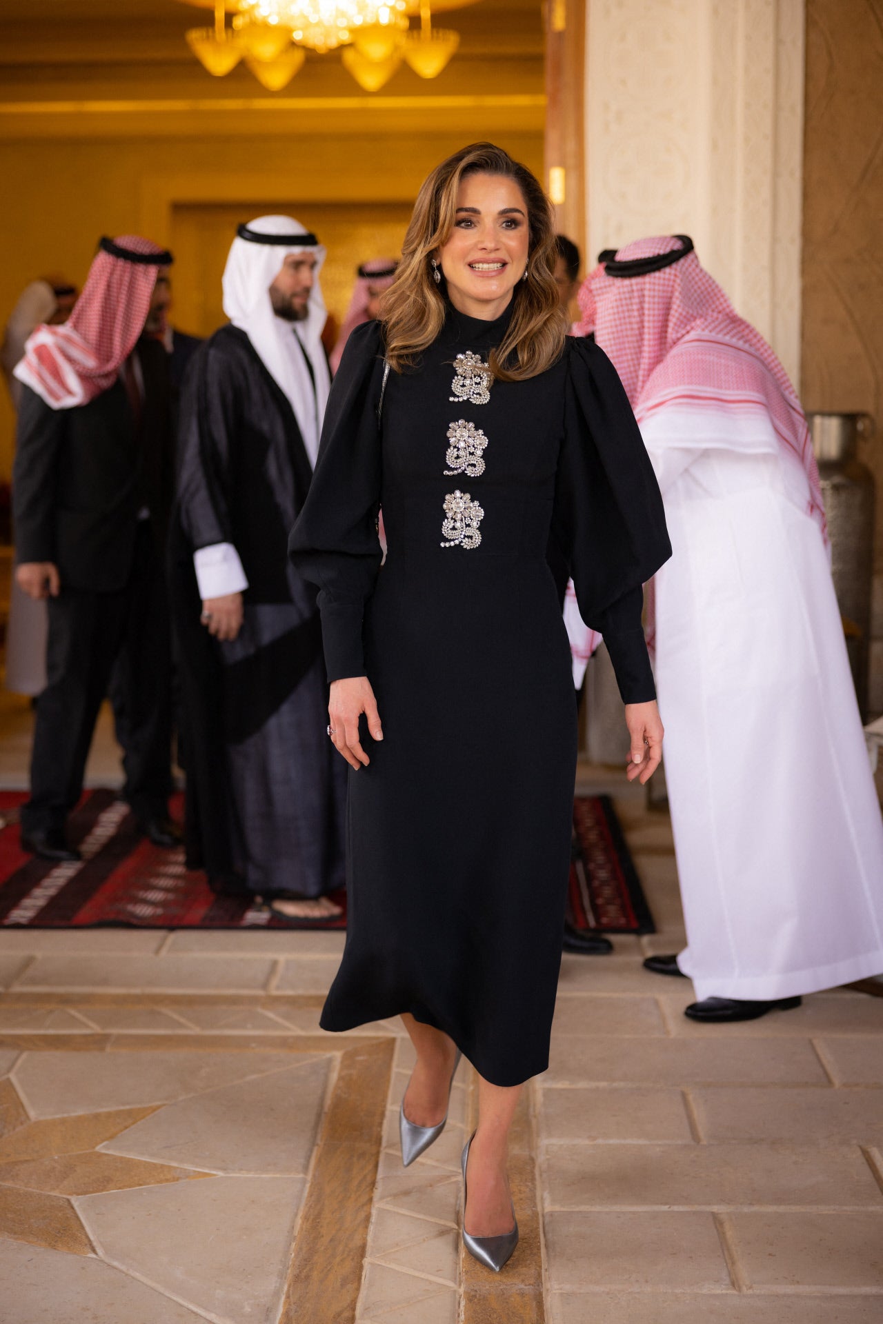 Para la celebración del compromiso de su hijo  Hussein, Rania eligió un sofisticado vestido negro de crepé que, al igual que el diseño que escogió para este día su futura nuera, consta de un cuello alto y mangas largas abullonadas, y pertenece al diseñador Andrew GN. El detalle que le aporta un plus de elegancia son unas  incrustaciones de cristales en forma de flor que hacen que la reina no necesitase llevar más joyas que unos pendientes y un anillo.