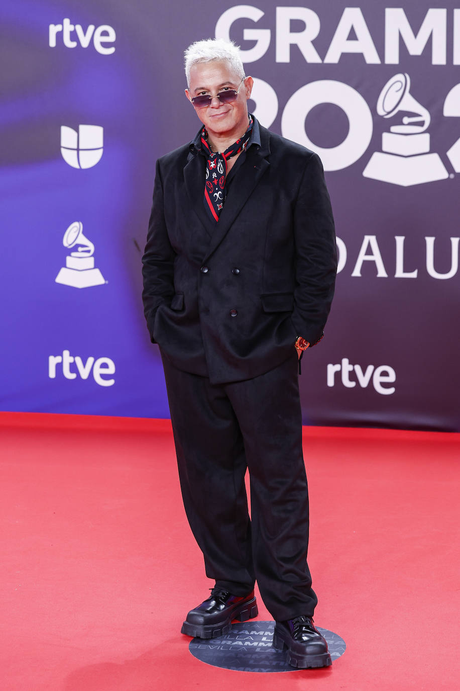 Alejandro Sanz con traje negro oversize en color negro de punto elástico de americana cruzada de doble botonadura y pantalón ancho combinado con un pañuelo en los colores insignia de la marca, todo de la colección Fall/Winter 2023 de HUGO.