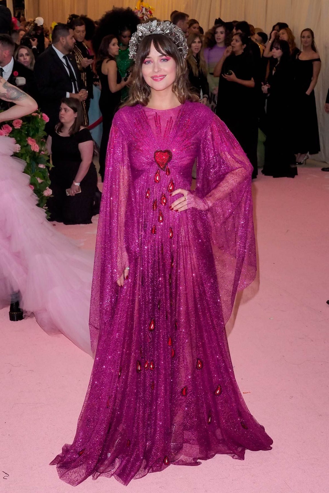 Elegante y sofisticada, Dakota Johnson llegó a la Gala Met de 2019 con un vestido largo con manga murciélago de color morado con microlentejuelas y decorado con un corazón rojo en el pecho del que caen gotas que simulan ser de sangre. Era de Gucci y lo completó con una diadema plateada.