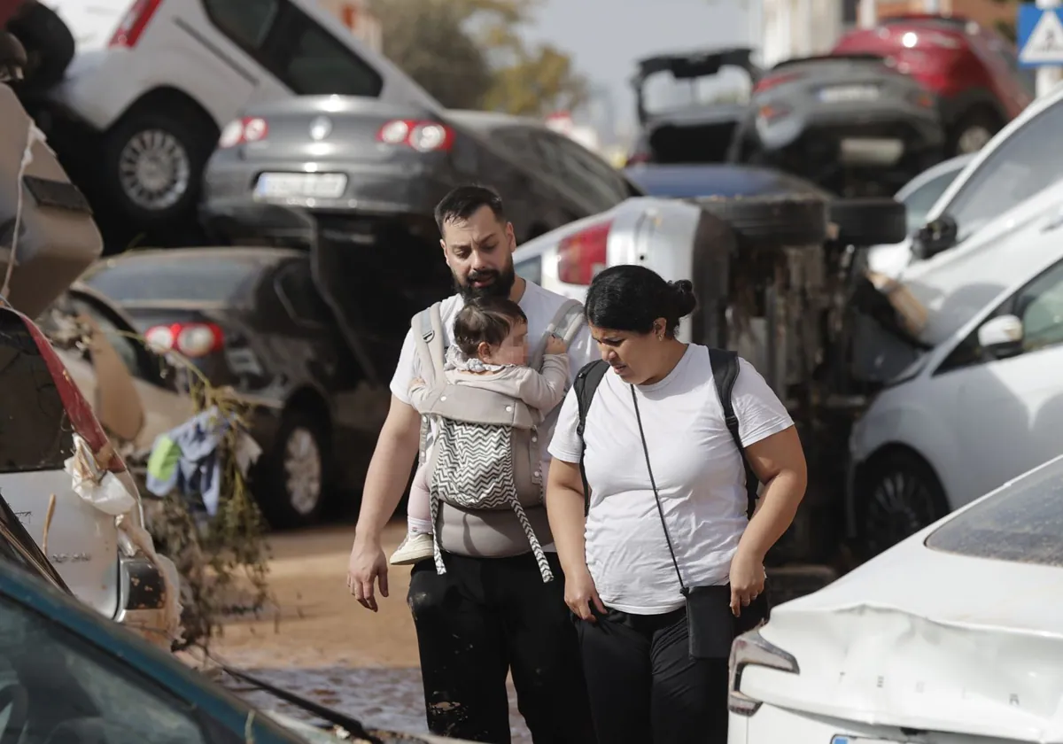 Una pareja con su bebé camina entre una montaña de coches hacinados tras las inundaciones sufridas en la localidad valenciana de Paiporta