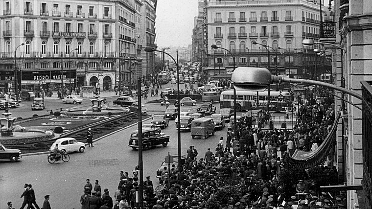 Ambiente de la Puerta del Sol durante el día de la Fiesta de la Banderita. Gran cantidad de público congregado en torno a la mesa instalada ante la dirección general de seguridad, junto a la banda de música