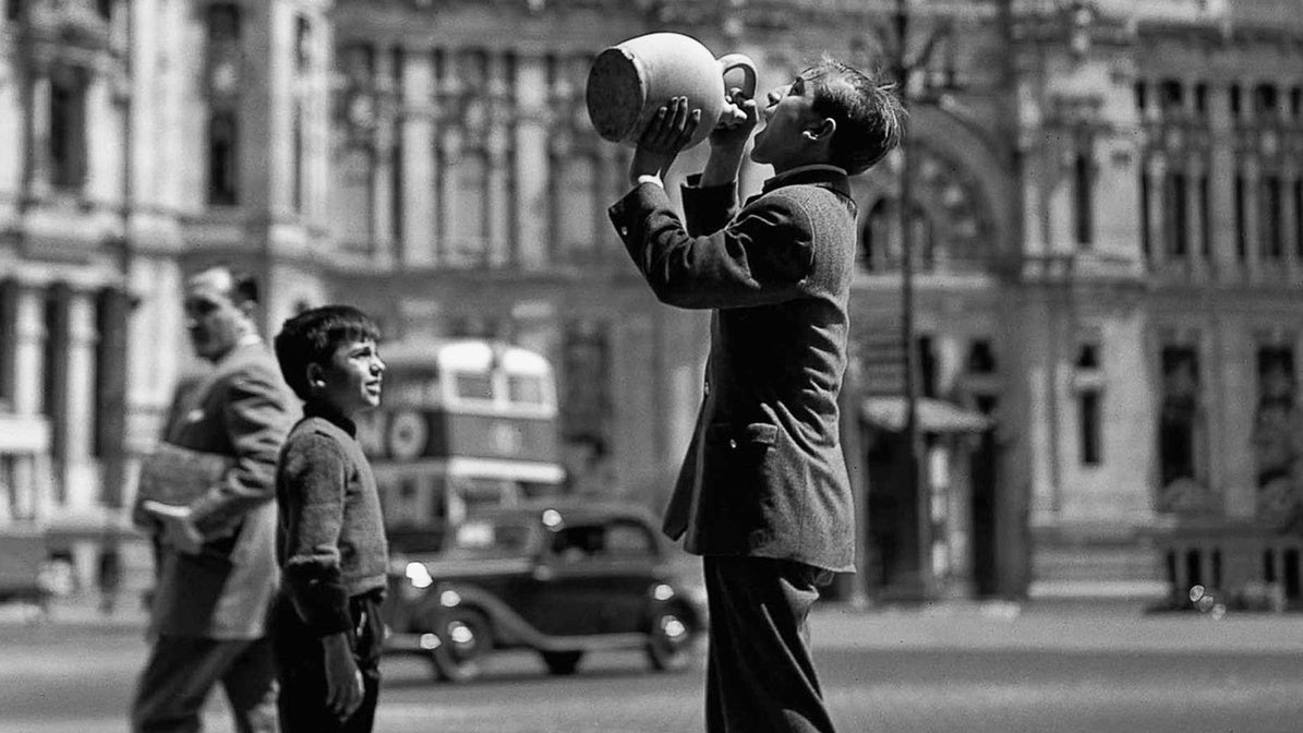Un adolescente bebe de un botijo frente al Palacio de Comunicaciones de Madrid, en 1952