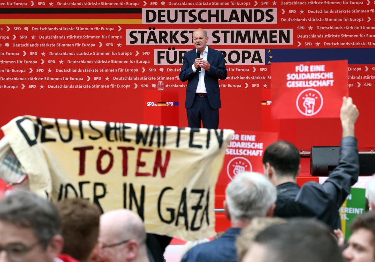 Olaf Scholz habla mientras un manifestante sostiene una pancarta que dice «las armas alemanas matan a niños en Gaza» durante la campaña electoral del SPD para las elecciones europeas