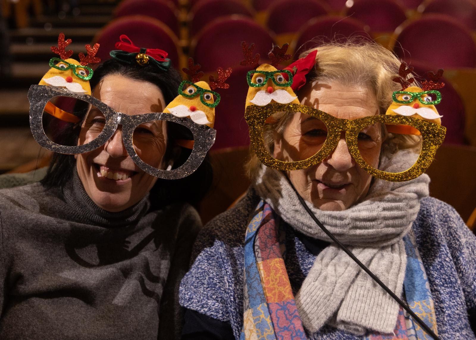 Dos mujeres, con gafas con motivos navideños, sentadas en el teatro.