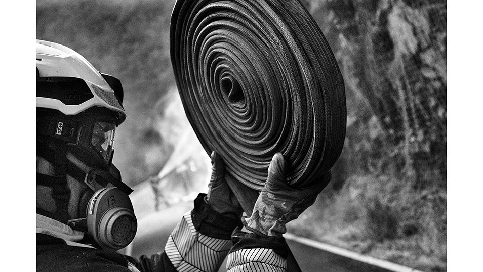 En primer plano, un bombero prepara una manguera para extenderla sobre el asfalto. Al fondo, entre el humo, otro compañero combate las llamas en la ladera. La escena resume la dureza del operativo en Fornela: mientras unos aseguran el material, otros se enfrentan directamente al fuego.
