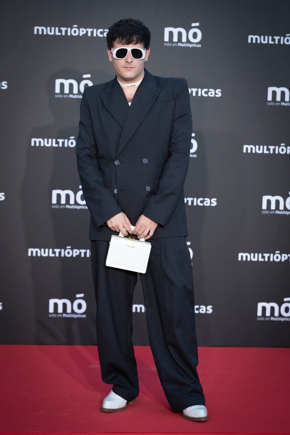 El diseñador de moda Palomo posa sobre la alfombra roja con traje negro cruzado y un minibolso blanco a juego con sus gafas de sol.