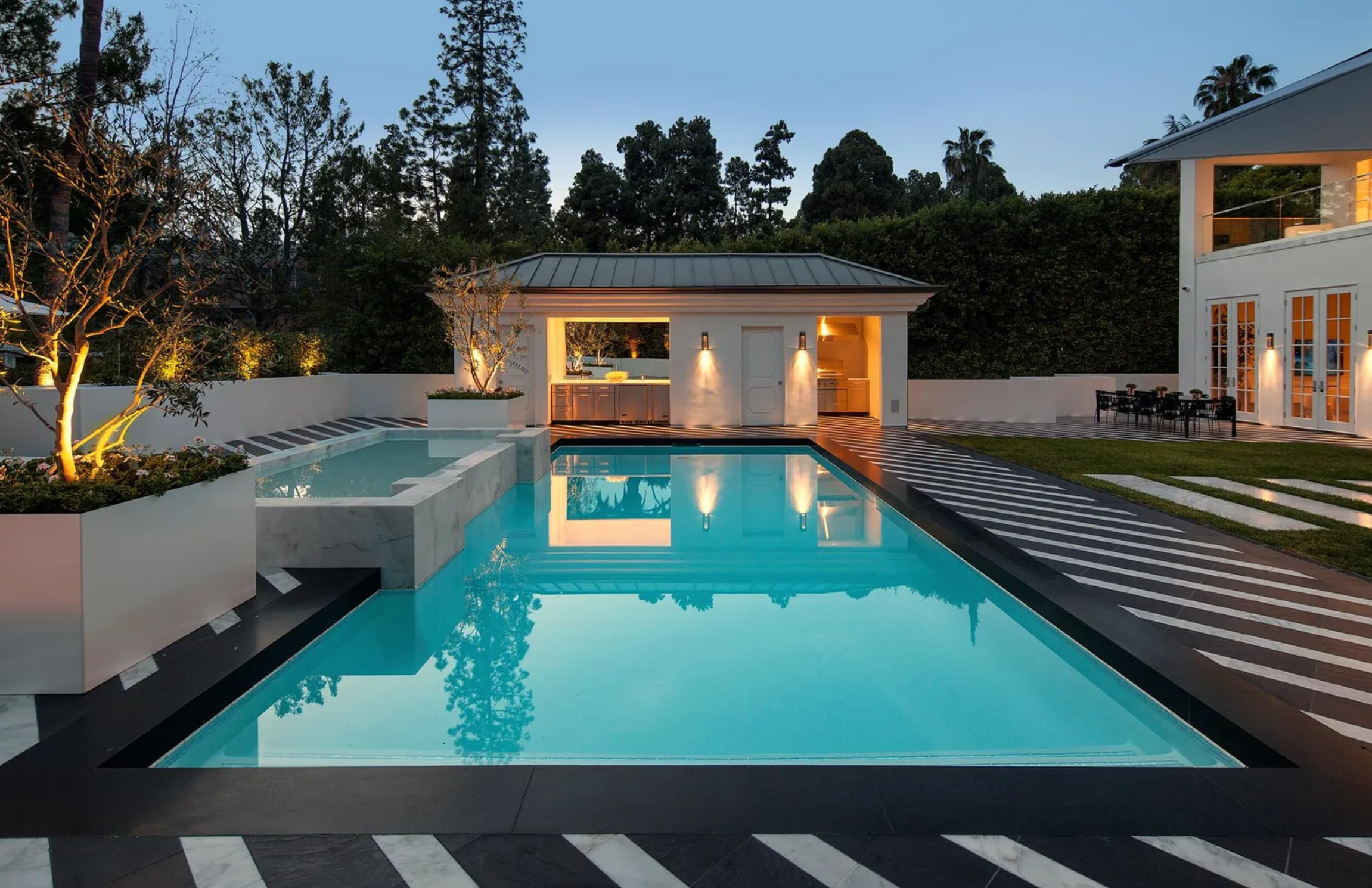 La zona exterior incluye una piscina con jacuzzi, cocina exterior, área ajardinada con grandes palmeras y un amplio garaje