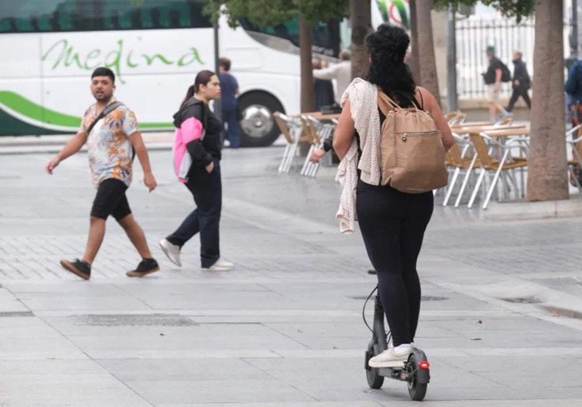 Una usuaria de patinete eléctrico pasa ante dos personas en la plaza de San Juan de Dios de Cádiz