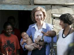 La Reina Sofía, sostiene dos bebés durante la visita que ha realizado durante su visita oficial, al barrio de Los Mina en Santo Domingo, a orillas del río Ozama. / Efe
