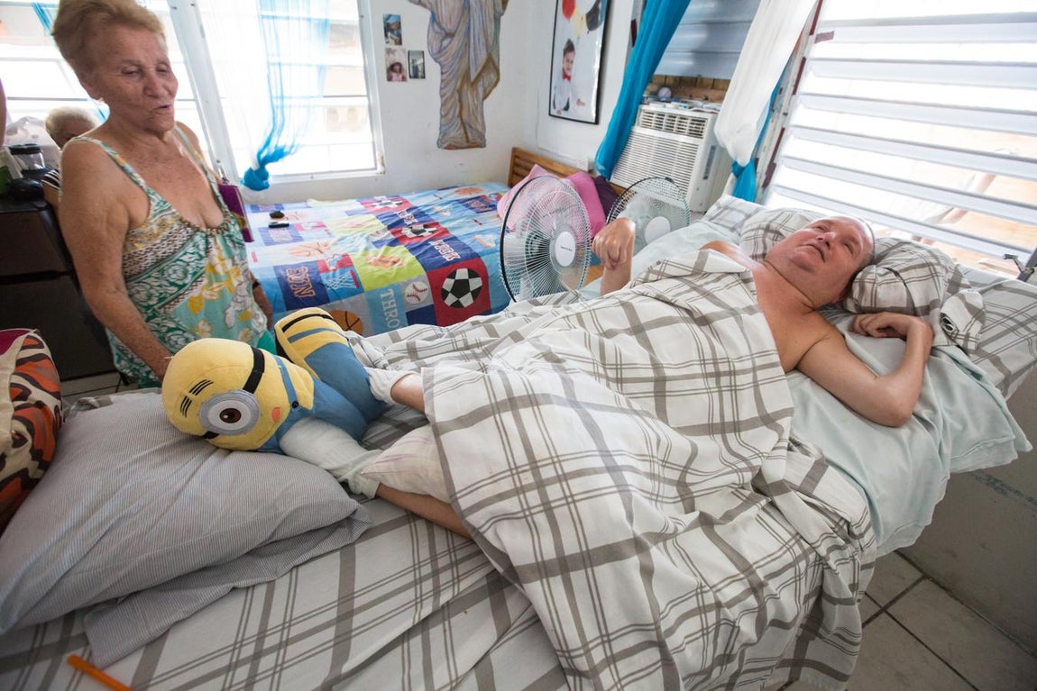 Jorge Carrasquillo es un ‘encamado’, como aquí llaman a la gente postrada en la cama. Sufrió un accidente en su juventud que le dejó inválido. En casa de su madre, como en el resto del barrio, no hay electricidad. Se alivia del calor con dos ventiladores que le apuntan a la cara, prendidos con un generador.. 