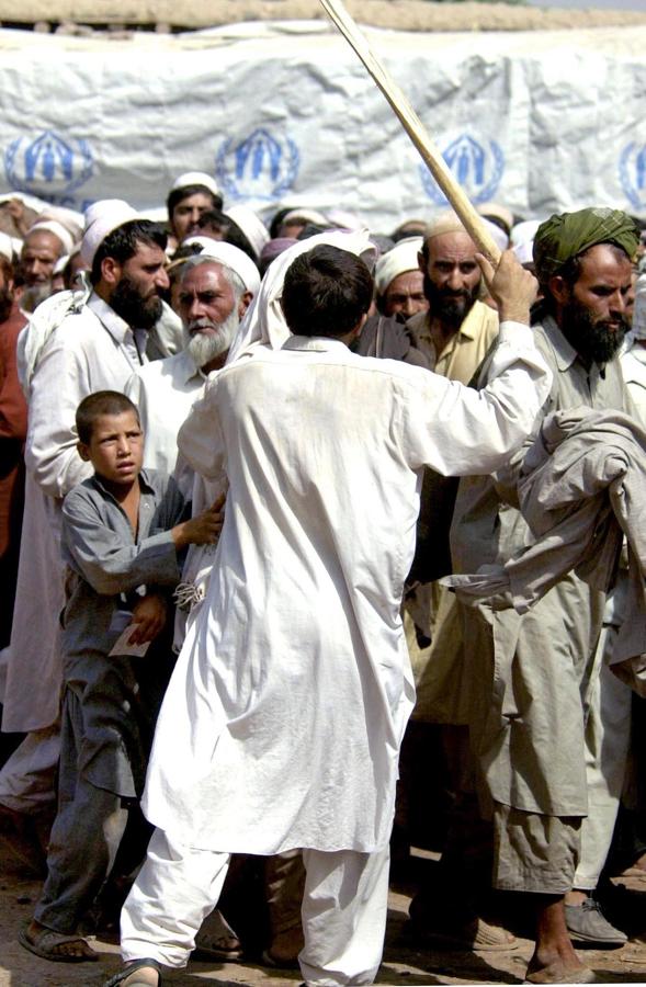 Varios afganos en un campo de refugiados, amenazados con un palo porque no guardan el orden en la fila para el reparto de la ayuda humanitaria