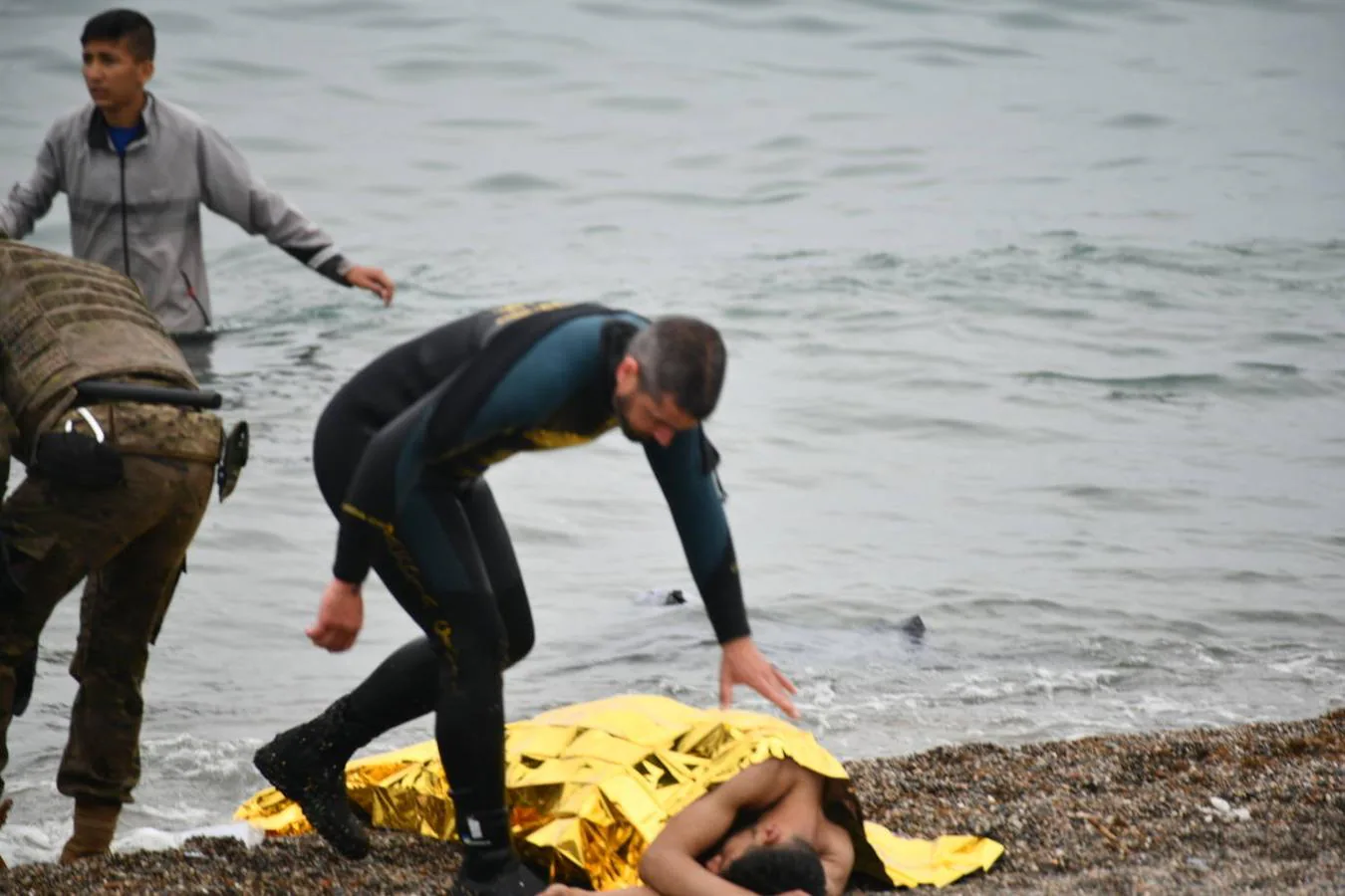 Un militar español ayuda a una persona tumbada en la orilla. 