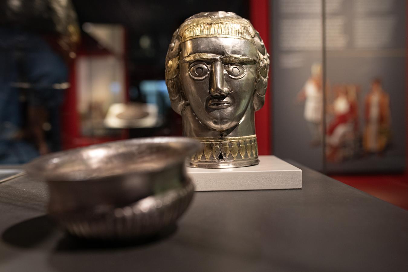 Vasija de plata, parcialmente dorada, con forma de cabeza de una mujer joven de pelo corto que lleva una diadema de plumas sobre la frente y un collar de cuentas en forma de anforetas. Muy probablemente, sea la representación de una deidad femenina. 