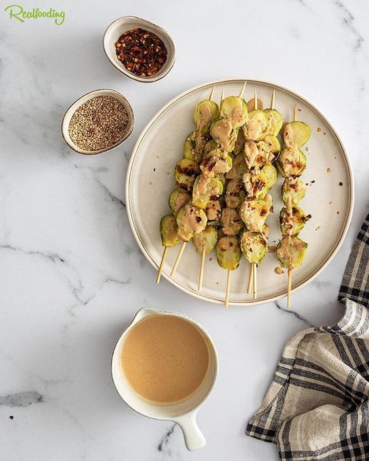 Brochetas de coles con salsa de cacahuetes. Cuece las coles de Bruselas durante 3 minutos en agua hirviendo con sal. Después corta las coles por la mitad y monta las brochetas. Pon una sartén al fuego con un poquito de aceite de oliva virgen extra y cocina las brochetas tres minutos por cada lado o hasta que estén ligeramente doradas. Para la salsa de cacahuete mezcla todos los ingredientes en un bol con un tenedor hasta que no queden grumos.