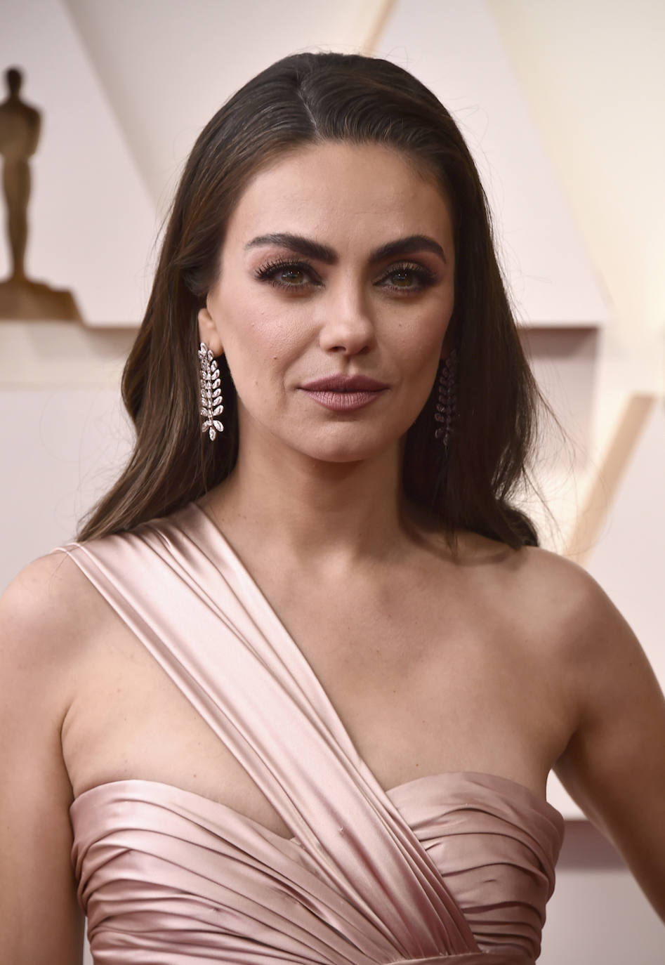 Mila Kunis con melena suelta, con raya a un lado, sombras de ojos en tonos tierra, máscara de pestañas y labios rosas en la alfombra roja de los Oscar 2022.