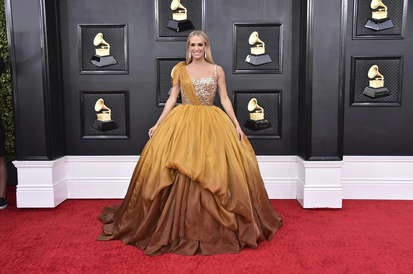 Carrie Underwood - Alfombra roja de los Grammy 2022. Cual princeda con un voluminoso vestido mostaza compuesto por un cuerpo ajustado con lentejuelas y una falda mostaza con degradado.