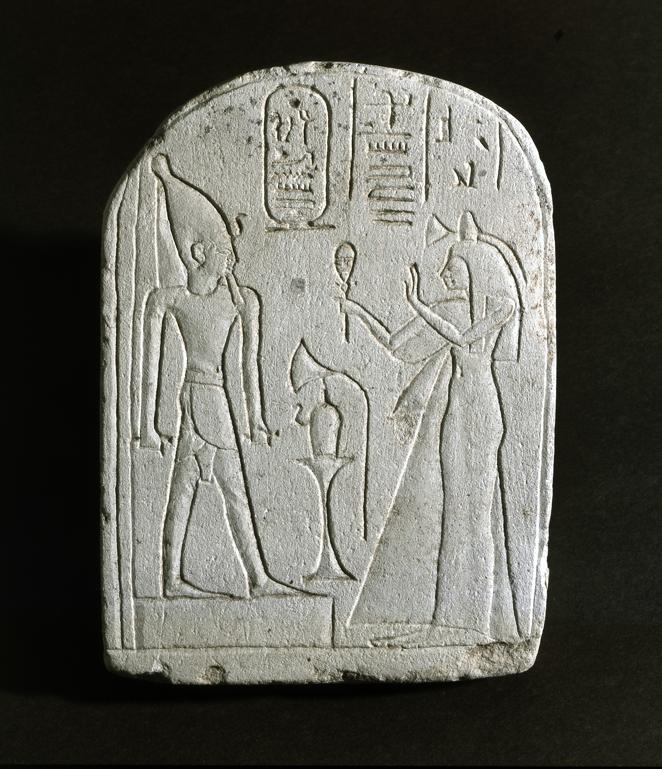 Isis ante el faraón. Estela del Reino Nuevo (1590-1070 a.C.) del museo de Hildesheim que representa a Isis, cantora de Montu-em-Taui sujetando un sistro con la mano derecha frente al faraón, con la corona del Alto Egipto. En la mesa de ofrendas representada entre ellos hay un vaso de libación y una flor de loto