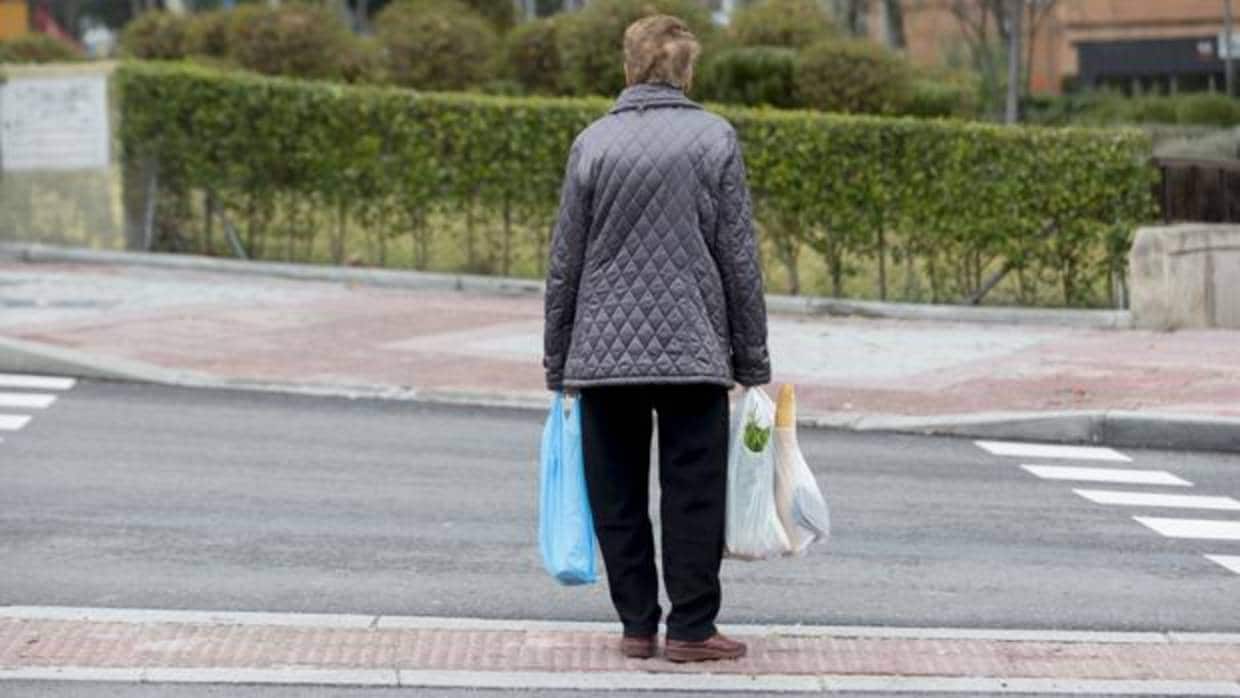 Una mujer de edad avanzada espera sola en un paso de peatones con bolsas de la compra