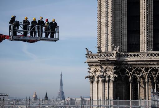 Seis trabajadores, en una grúa, junto a Notre Dame, con la Torre Eiffel al fondo