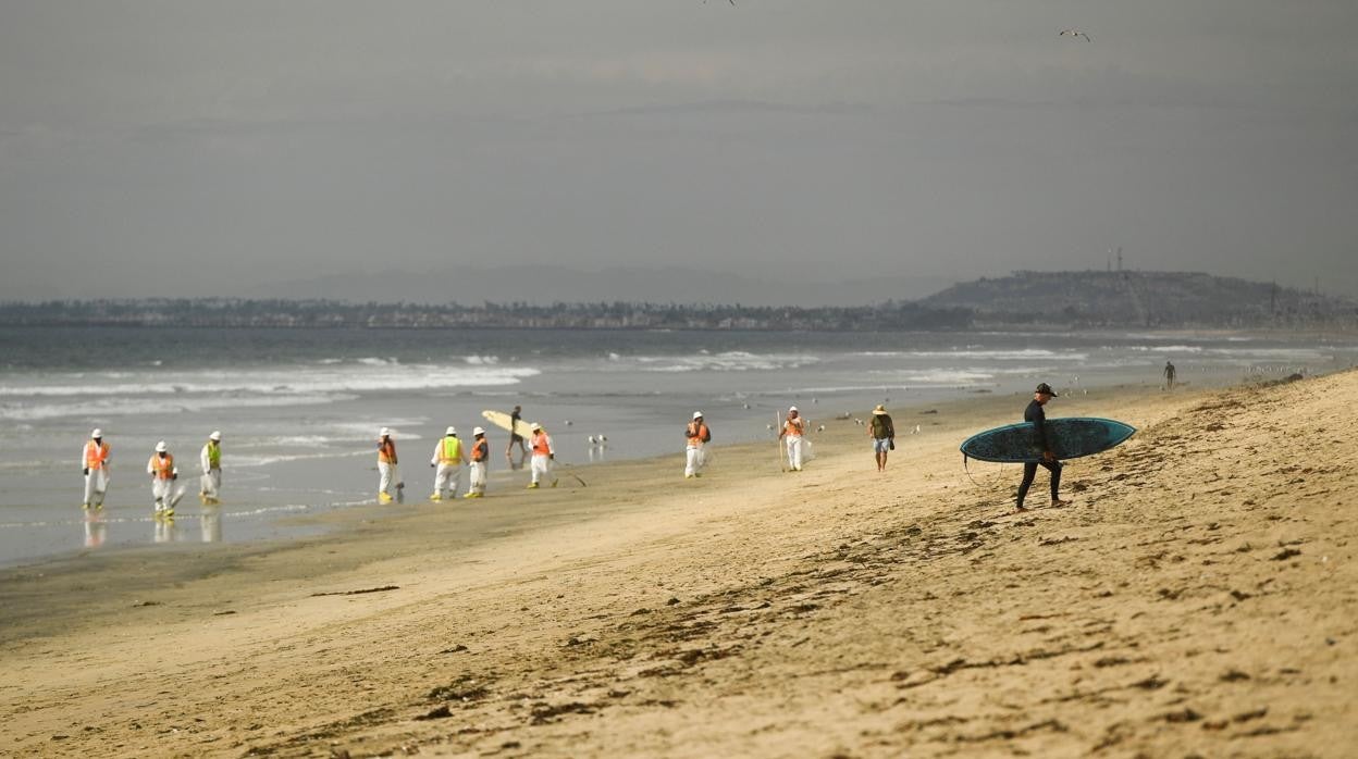 Un surfista abandona la playa de Huntington Beach mientras losoperarios limpian la playa