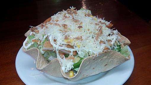 Ensalada Cesar, servida en un cesto crujiente de trigo, elaborada con hojas verdes de lechuga, pollo empanado, tomate, maíz y cebolla frita, adornada con queso rayado, y aderezada con nuestra salsa Cesar