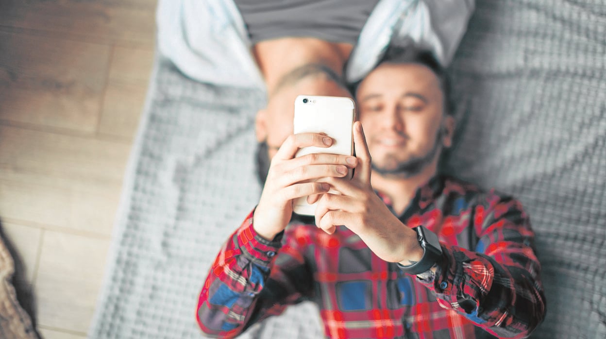 Un hombre tumbado en la cama, junto a otro, mira su móvil