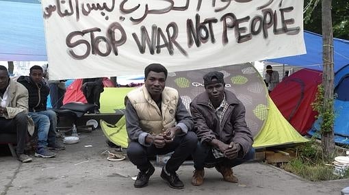 Dos refugiados en el campamento levantado en los Jardines d'Eole, con una pancarta que pide que paren las guerra pero que no se pare a la gente