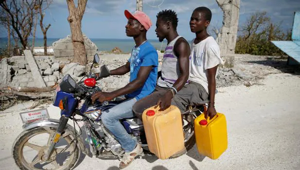 Tres Haitianos portan agua a bordo de una motocicleta frente a las ruinas de una casa derribada por «Matthew»