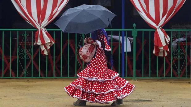 Una joven ataviada con el traje de flamenca se resguarda de la lluvia bajo un paraguas