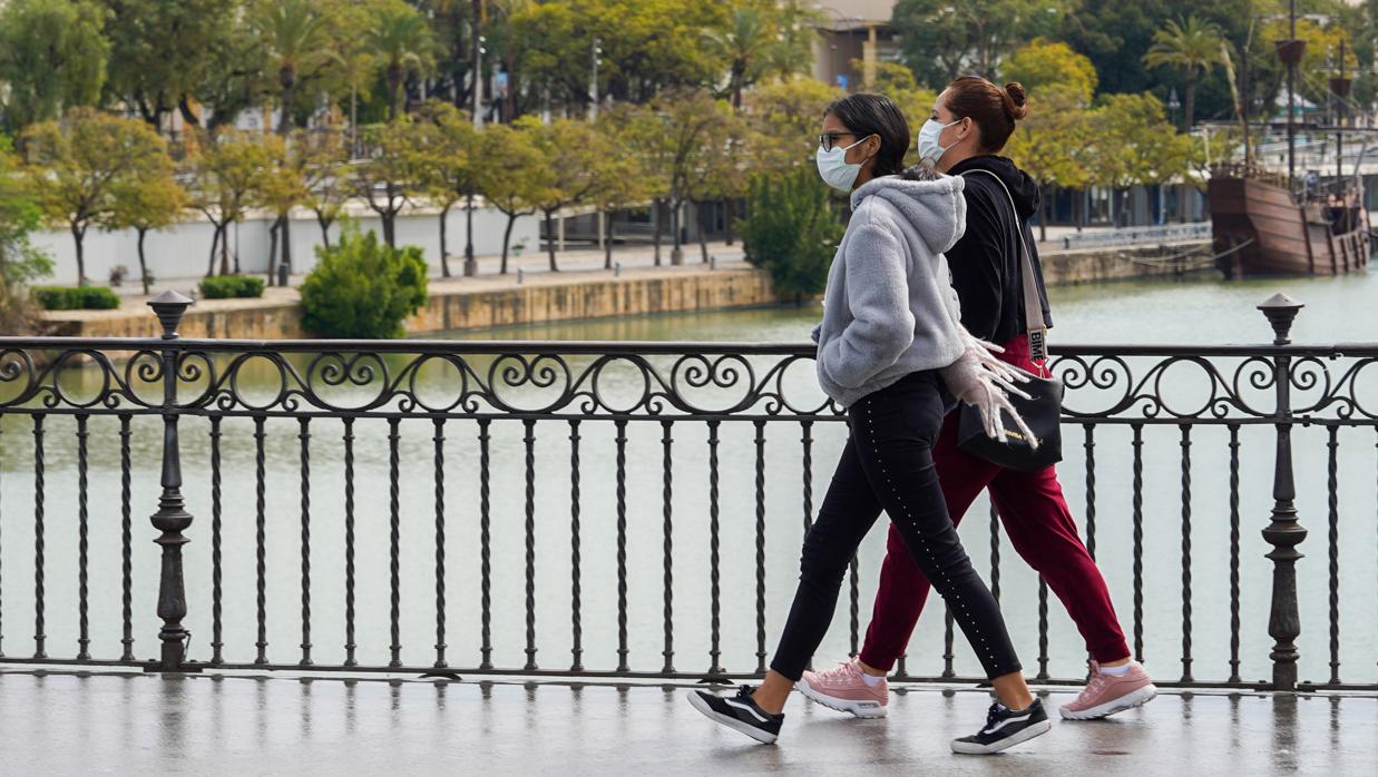 Dos jóvenes con mascarillas pasean andando por el puente de Triana