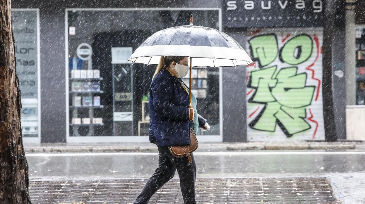 Una mujer pasea por Sevilla, protegida por una mascarilla y un paraguas, bajo la lluvia