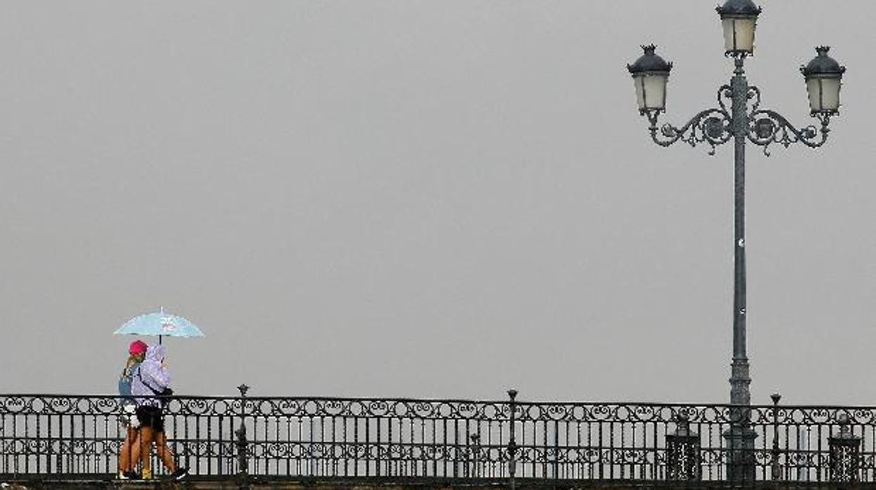 Un par de personas pasean por el puente de Triana protegidas de la lluvia con un paraguas