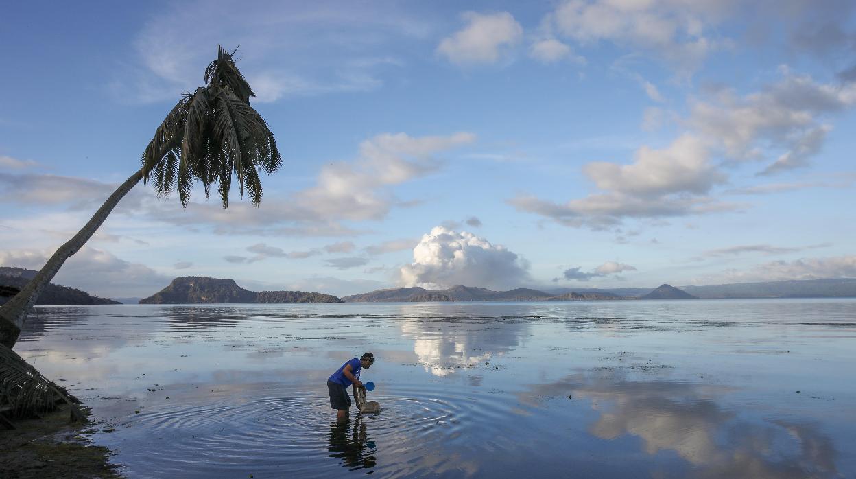 Un hombre lava sus utensilios llenos de ceniza en un lago de Tanuan (Filipinas) con el volcán Taal de fondo