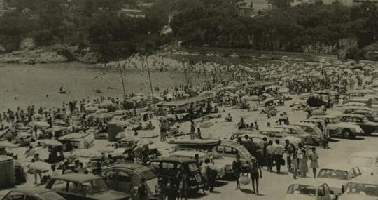 El aspecto en 1969 de la playa de Sant Feliu de Guíxols en agosto, repleto de bañistas que combatían el calor. Destaca el poco espacio que había entre la orilla y el aparcamiento
